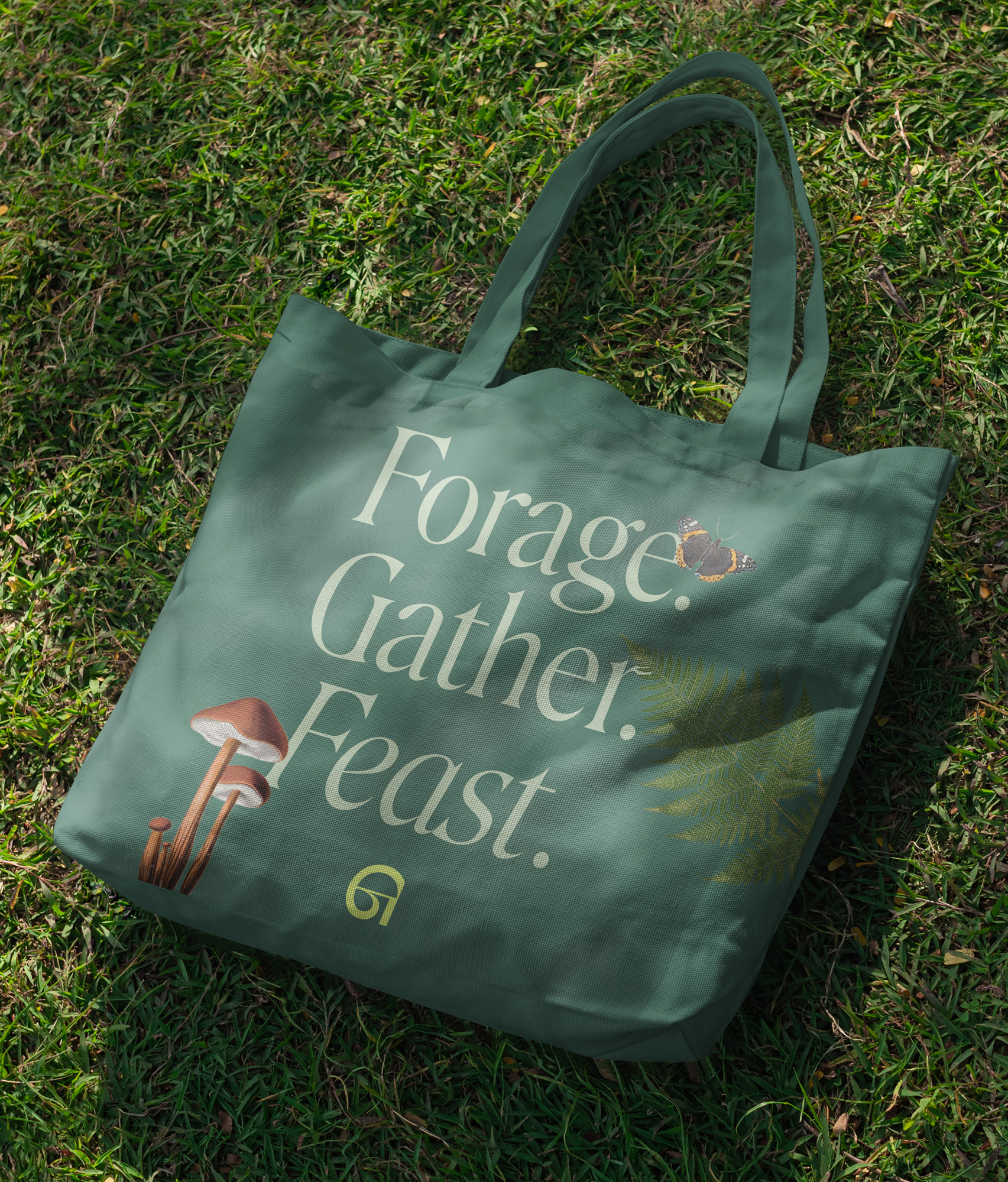 A green canvas tote bag lying on grass, collaged with mushrooms, a butterfly and a fern, with the Gather mark in yellow and the words Forage. Gather. Feast.