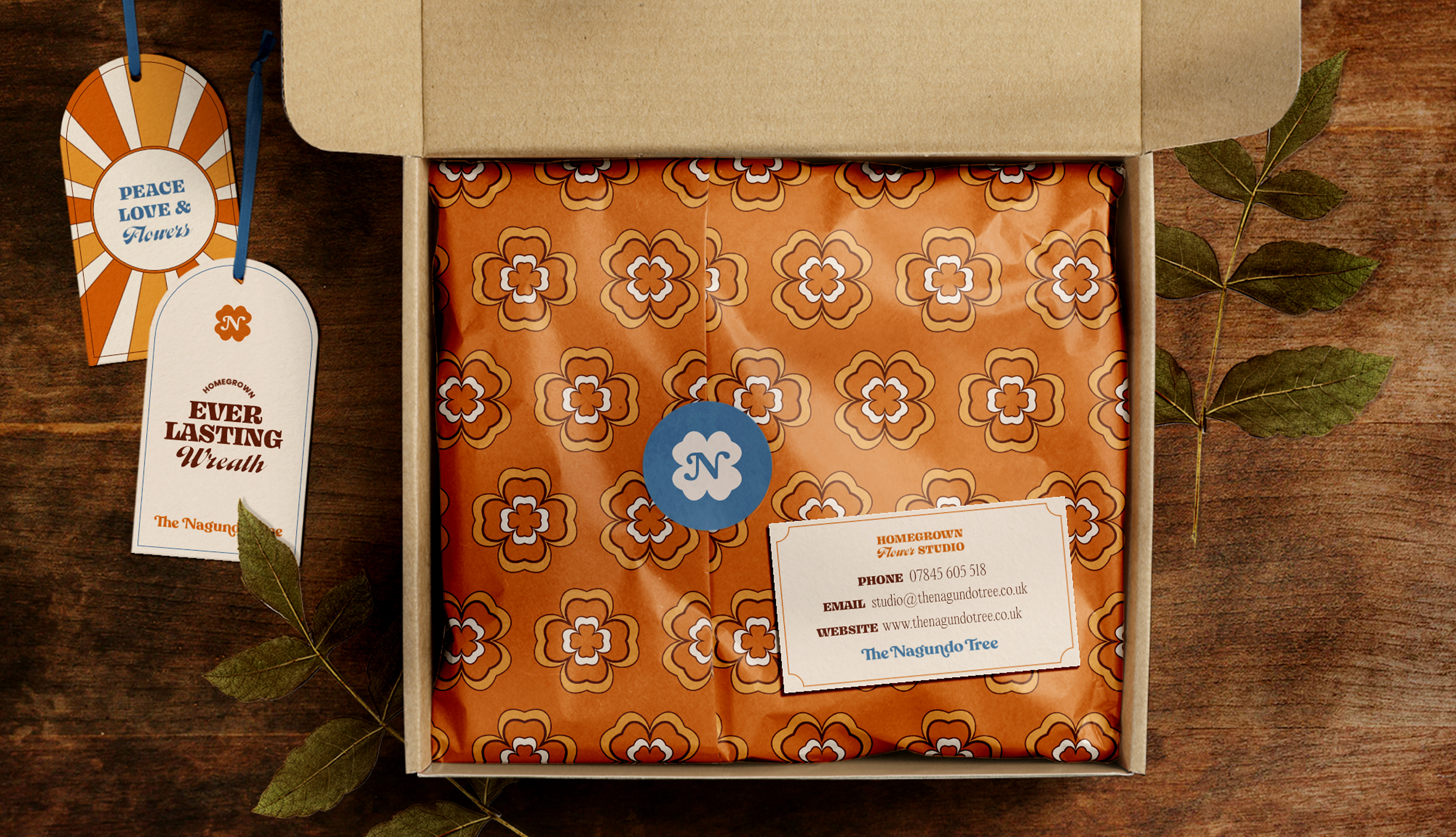 An open postal box of patterned tissue sealed with a clover sticker, with an Everlasting Wreath swing tag and a sunburst Peace, Love & Flowers tag alongside