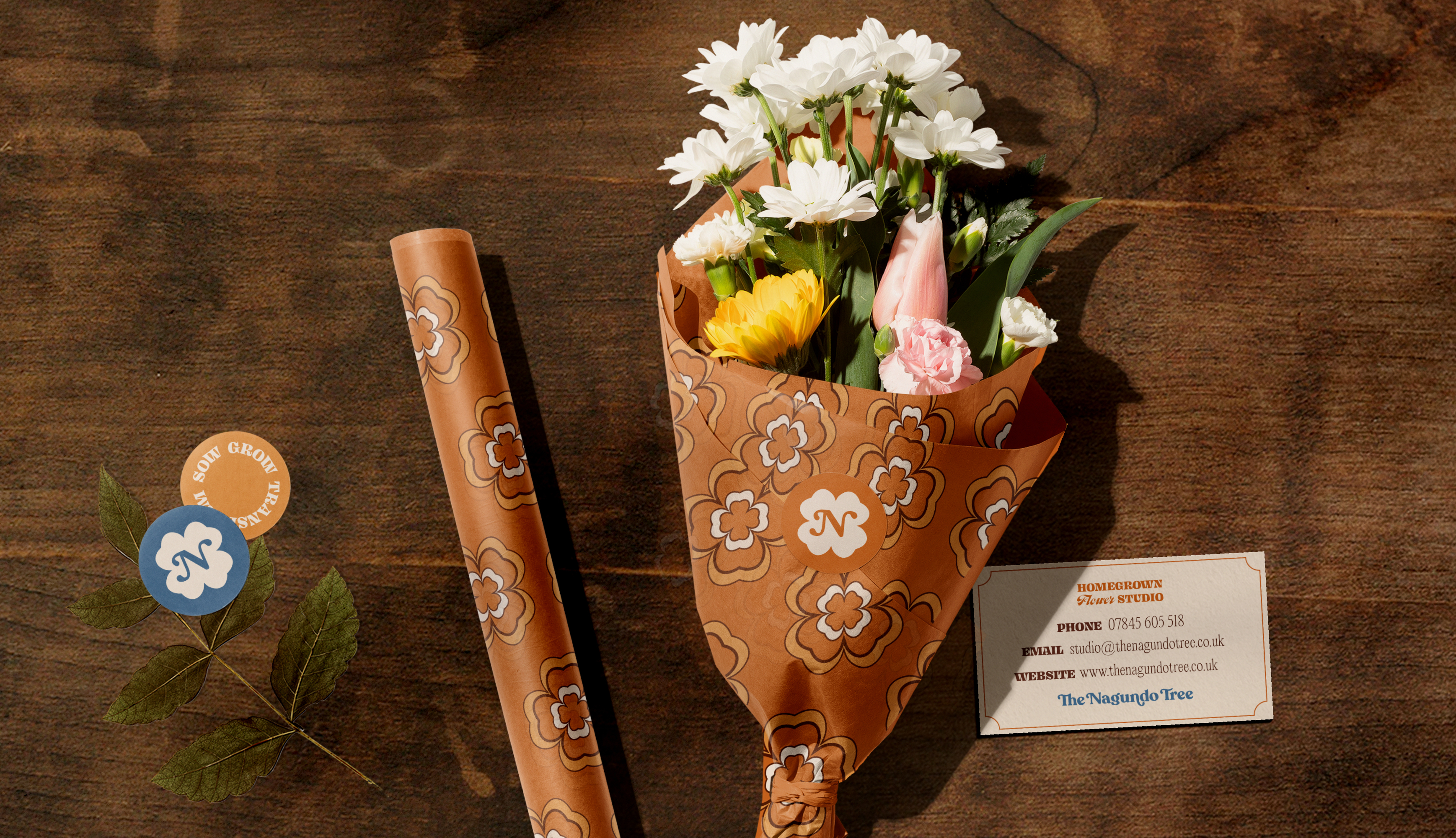 A bouquet wrapped in patterned paper and sealed with a clover sticker, beside a roll of the same paper and a business card