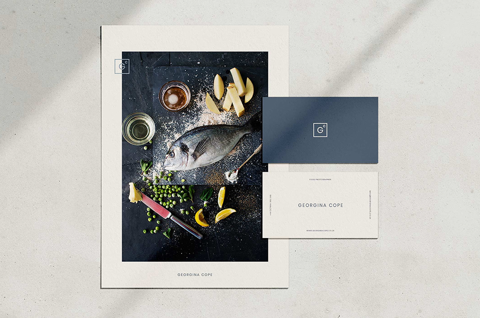 Georgina Cope stationery on a pale surface: a poster of an overhead food photograph of sea bream with peas and lemon, beside two business cards carrying the boxed GC mark