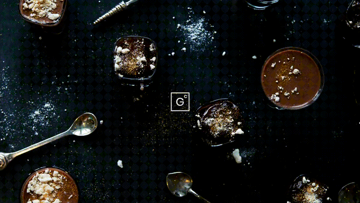 An overhead shot of chocolate pots dusted with crumb and spoons on a dark patterned cloth, the boxed GC mark centred over it