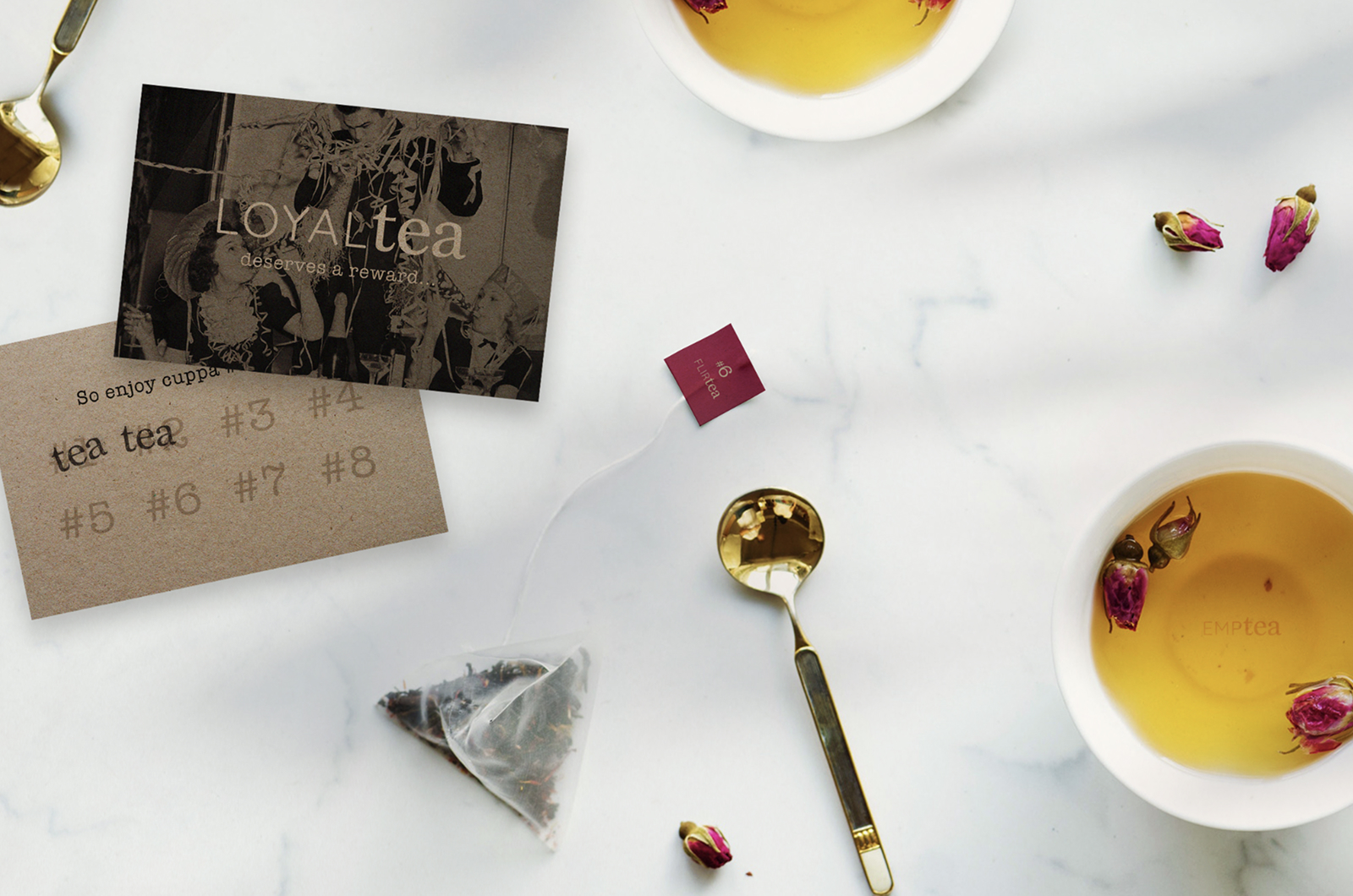 A LOYALtea card, a stamp card, a pyramid tea bag and gold spoons laid on marble beside two cups of rose tea