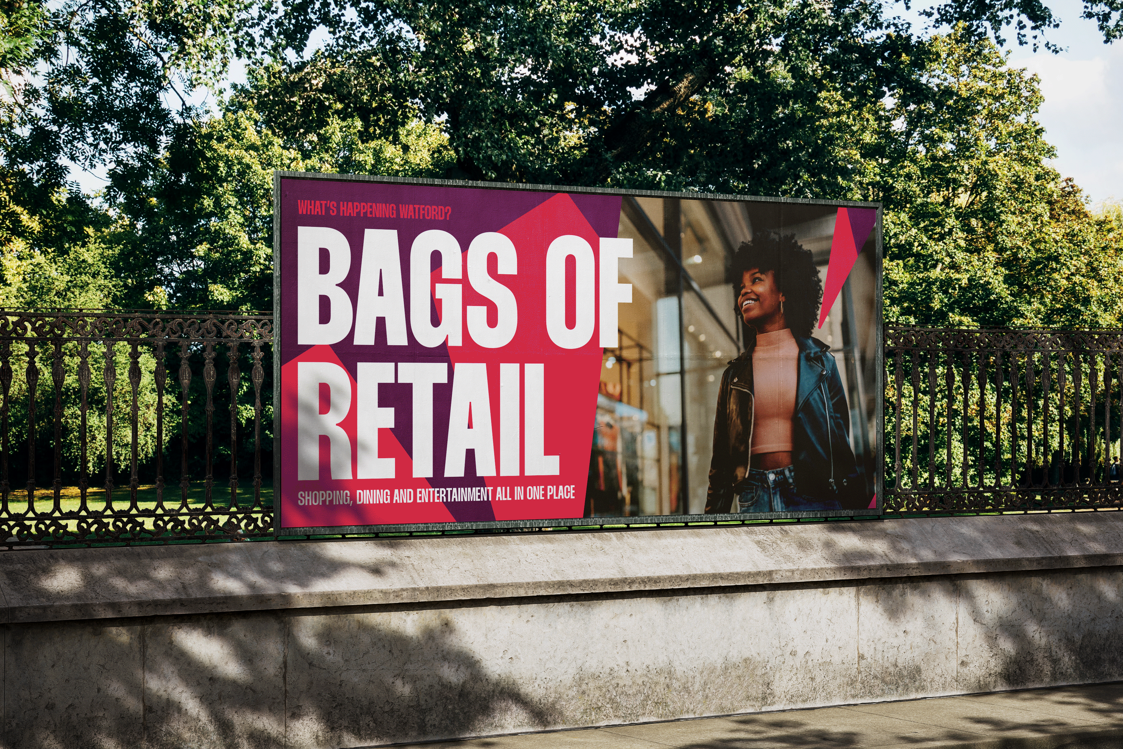 A billboard on a park railing reading bags of retail, set in heavy condensed type on magenta beside a shopper, under the line what's happening Watford