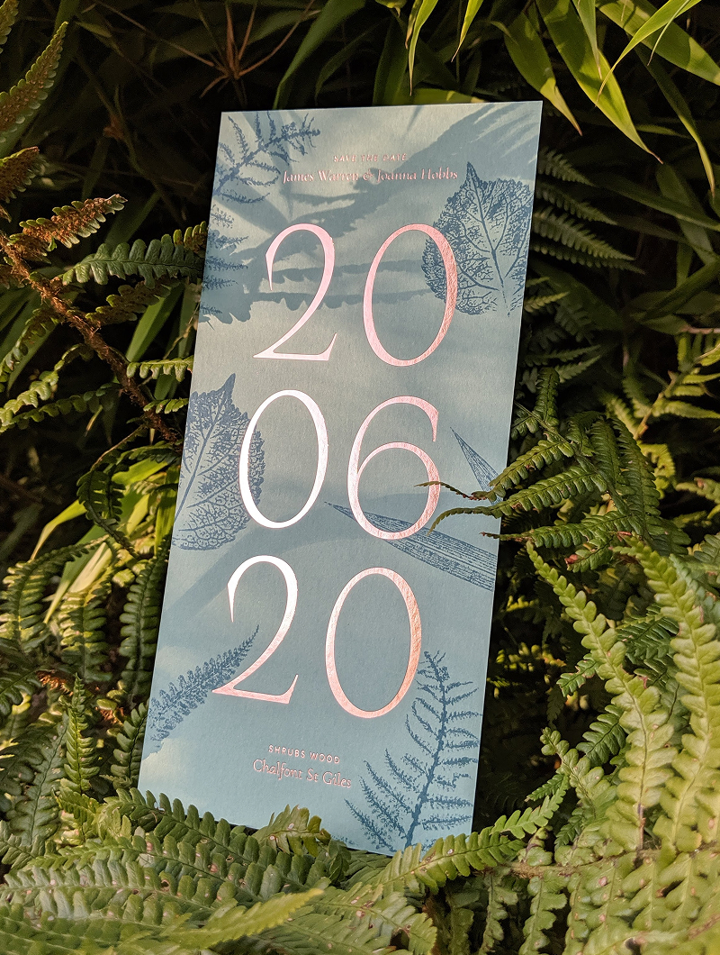A save the date card propped among ferns, the date set in large pink foil numerals over a blue ground printed with leaf prints