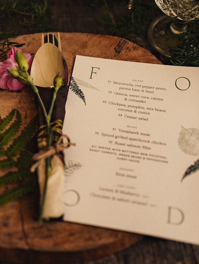 A wedding menu on a wooden board beside wrapped cutlery and a pink carnation, its courses framed by the letters F O O D and fern prints