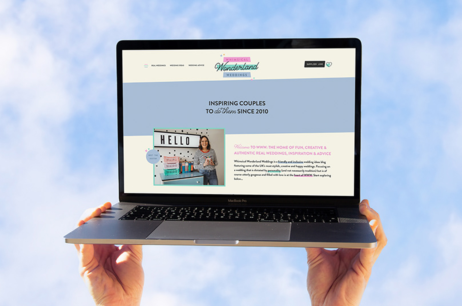 Hands holding up a laptop against a bright cloudy sky, its screen showing the Whimsical Wonderland Weddings homepage with the script logo and the line inspiring couples to do them since 2010