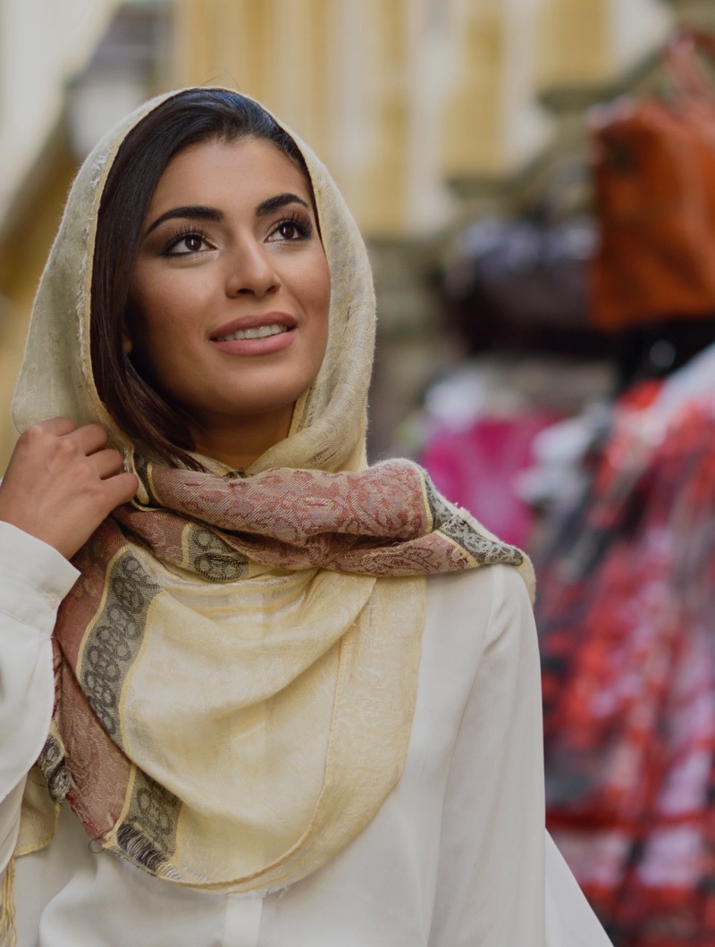 A woman in a cream headscarf looking up as she passes a market stall hung with leather bags