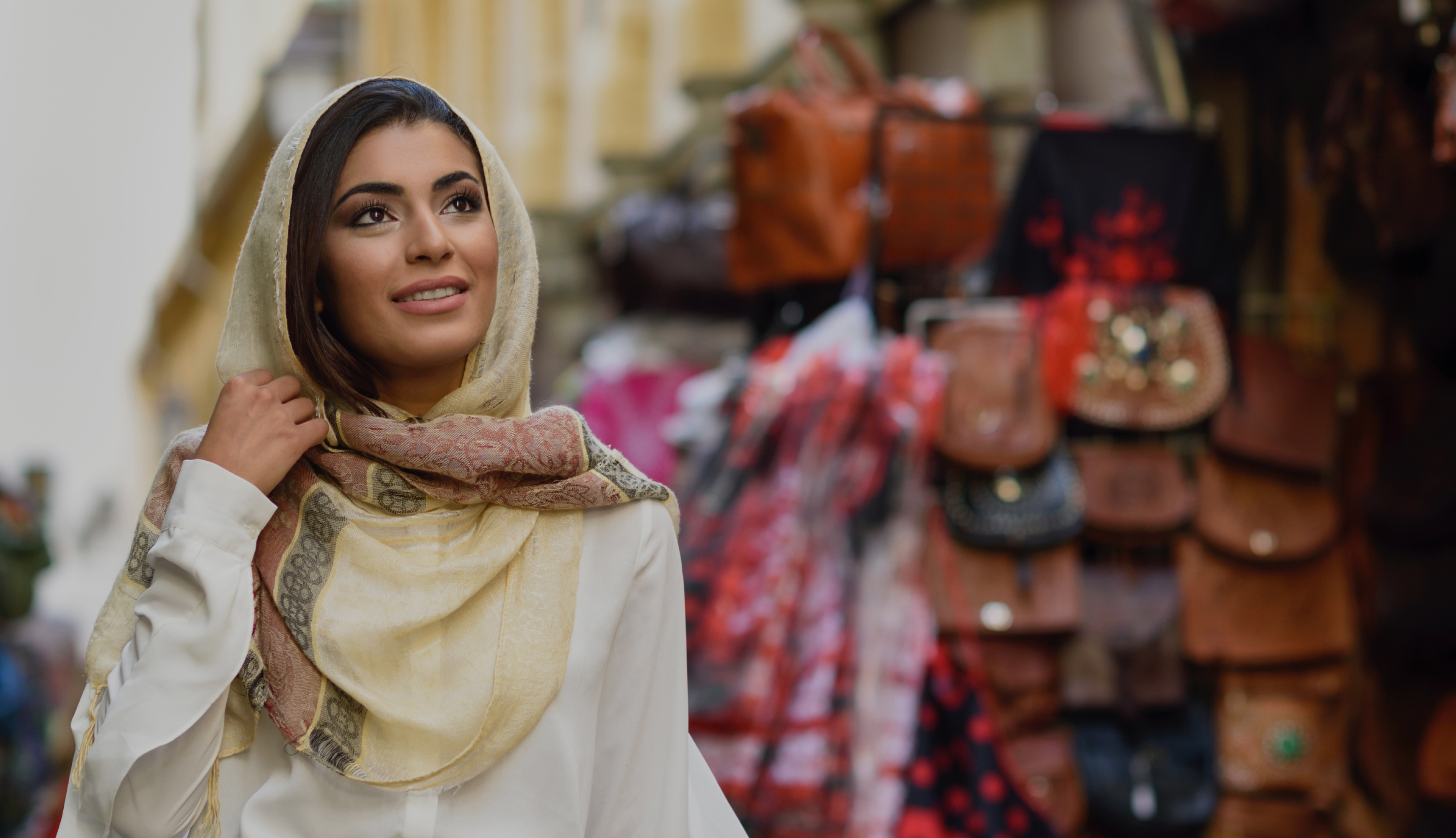 A woman in a cream headscarf looking up as she passes a market stall hung with leather bags