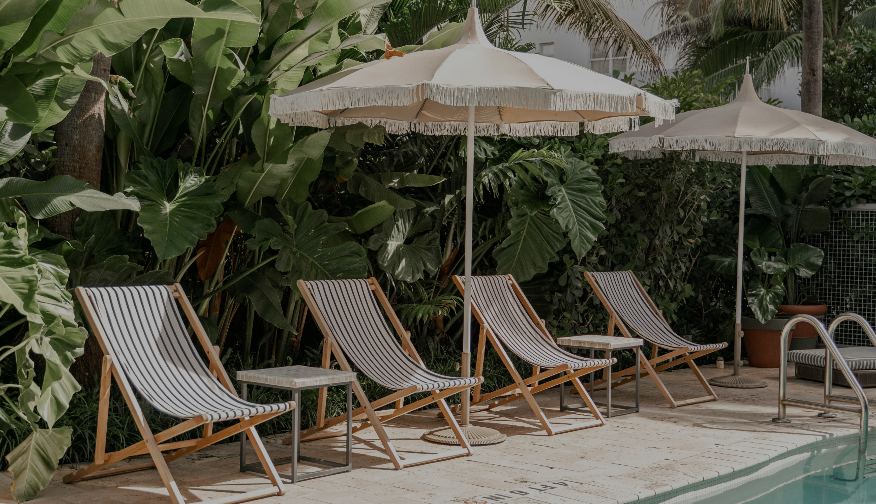 Striped deck chairs under fringed parasols beside a pool, backed by tropical planting