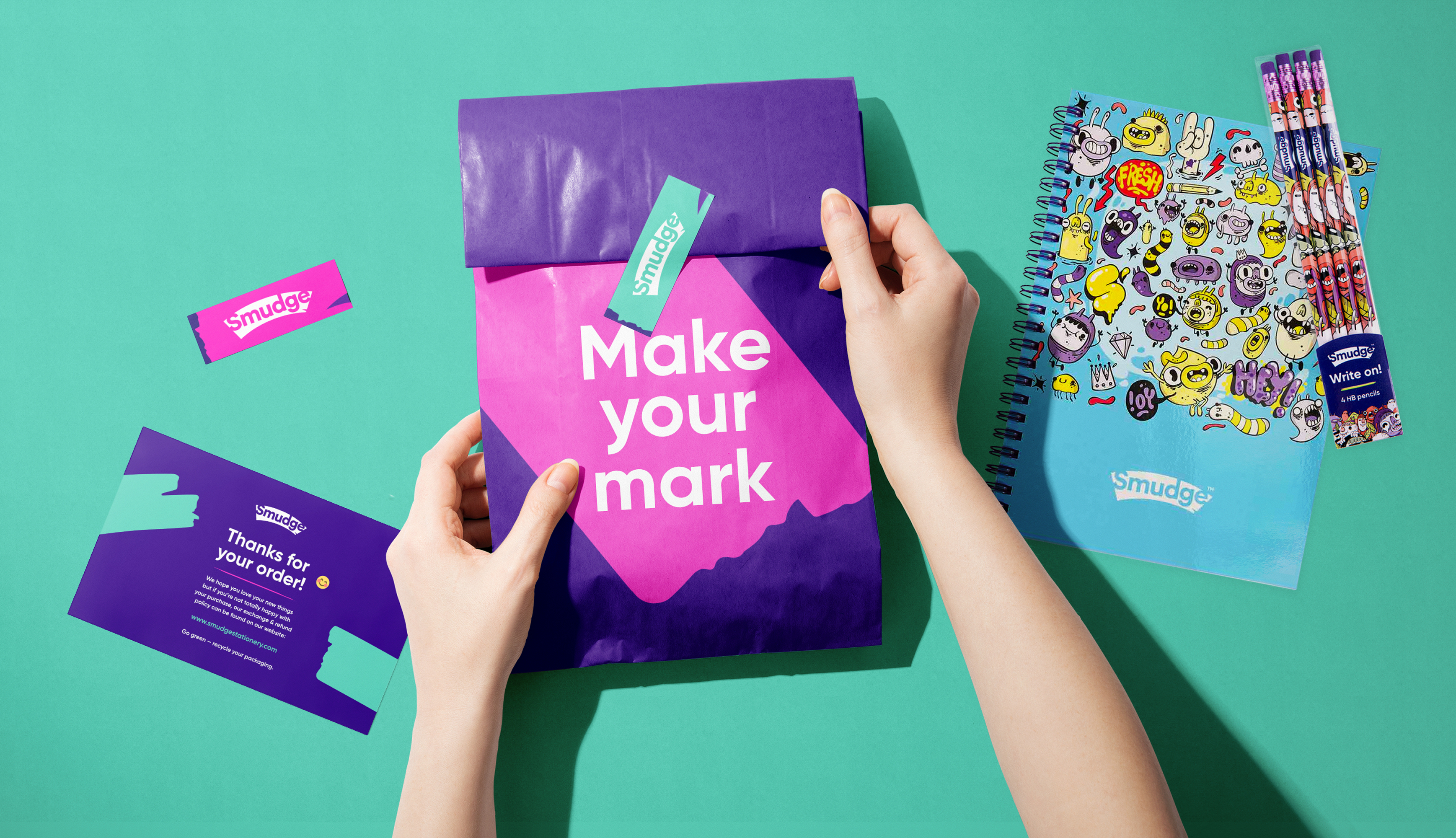 Hands holding a purple and pink Smudge mailer printed ‘Make your mark’, with a thank-you card, a branded sticker and a doodled notebook on mint green